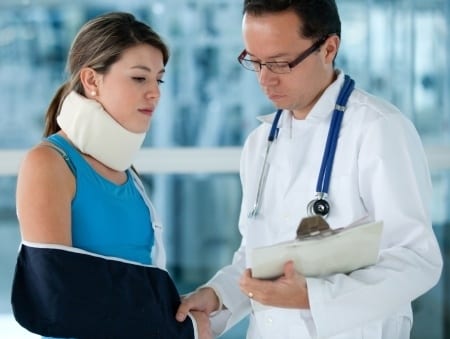 A woman with a neck brace and arm sling stands next to a male doctor holding a clipboard. The doctor is wearing glasses, a white coat, and a stethoscope. They appear to be discussing something in a medical setting.