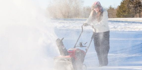 A person wearing a winter coat and hat operates a snowblower in a snowy landscape. The machine is blowing snow to the side. Trees are visible in the background under a clear sky.