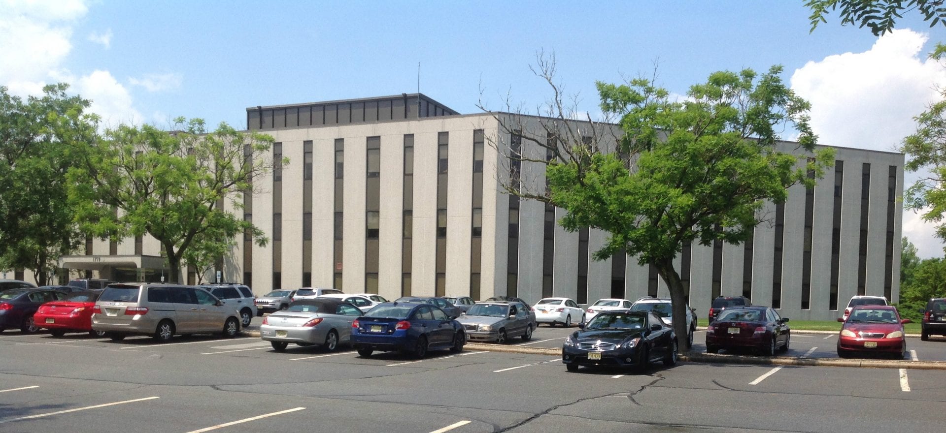 A large, rectangular office building with vertical windows stands behind a parking lot filled with various cars. Tall trees partially shade the area. The sky is clear and blue.