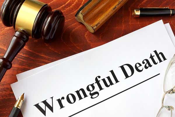 A wooden gavel rests on a desk next to a piece of paper with the words "Wrongful Death" in bold black letters. Nearby are a pen and a pair of glasses, suggesting a legal setting.