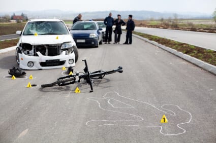 A car with a damaged front is stopped on a road next to a fallen bicycle and a chalk outline. Several markers are placed around the scene, and four people are talking in the background.