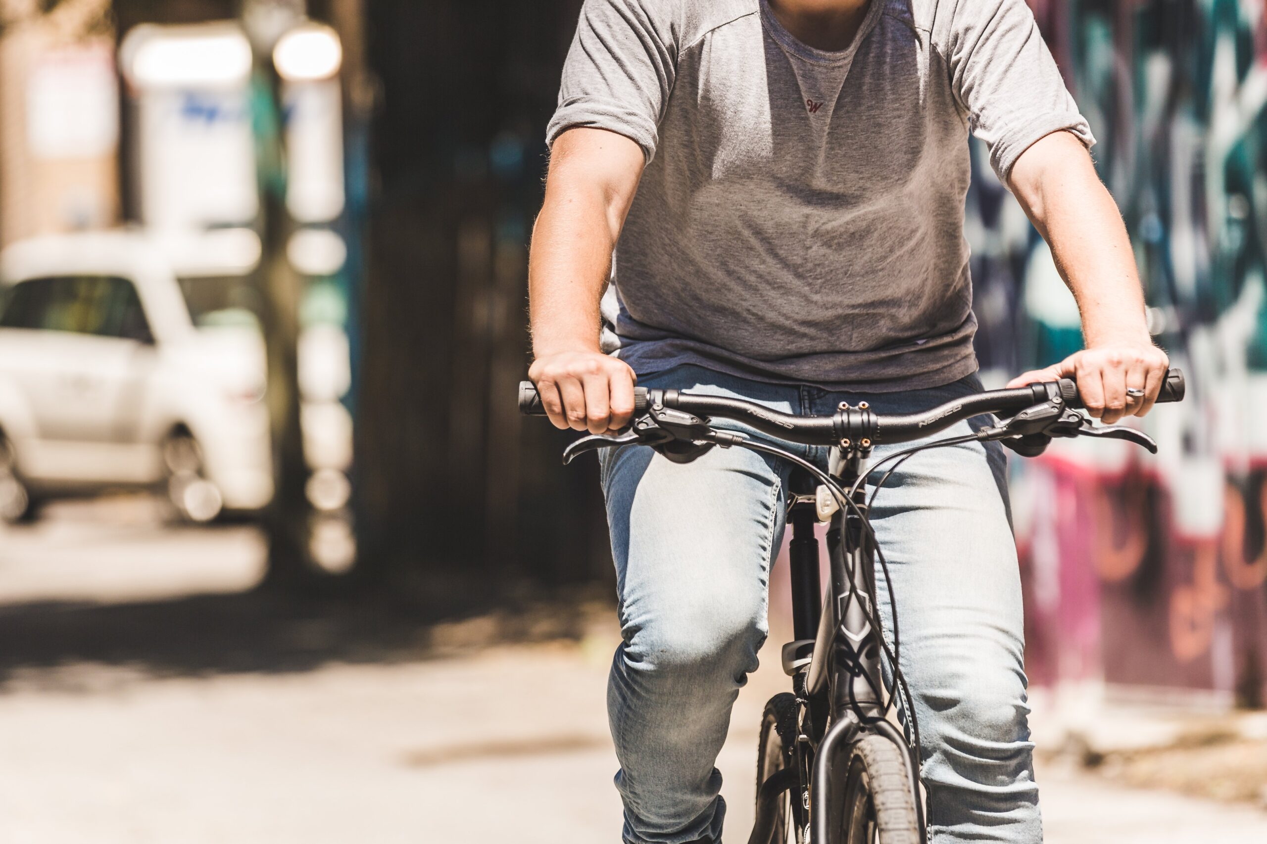 A person in a gray shirt and jeans rides a black bicycle through an urban area, passing a white car and a graffiti-covered wall with blurred buildings. The scene captures the vitality and energy of city life—something bicycle accident attorneys know too well.