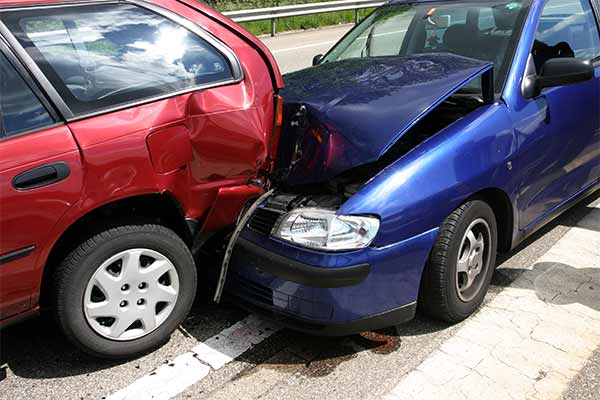 A red car and a blue car are involved in a rear-end collision on the road. The front of the blue car is damaged and crumpled against the back of the red car, which has a noticeable dent. The scene is during the daytime.