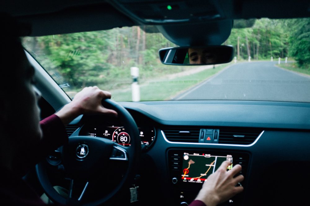 A person is driving a car on a narrow road surrounded by greenery, with the speedometer reading 80. They check the touchscreen navigation system frequently, aware of how critical it is to drive safely—knowledge perhaps courtesy of advice from car accident attorneys. The rearview mirror reflects part of their face.