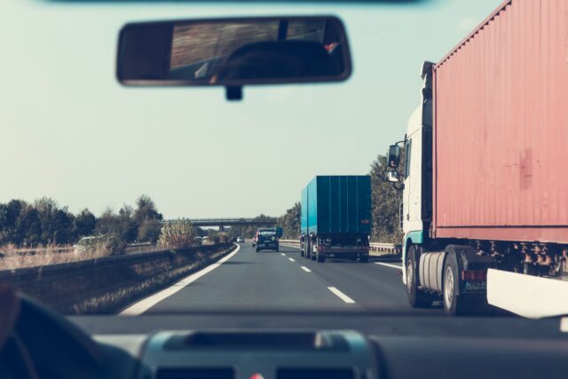 From inside the car, the expansive New Jersey highway scene unfolds with two large trucks carrying containers and a smaller vehicle up ahead. The road leads toward an overpass in the distance, framed by trees under a clear sky—a peaceful drive unmarred by any truck accident concerns.