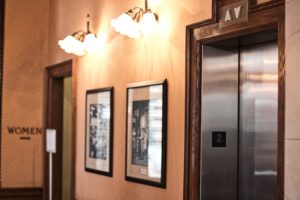 An elevator with a wooden frame displays the number two above its doors. Two framed pictures adorn the adjacent wall, while "Women" is visible near a door. Understanding safety features can help prevent accidents, a crucial consideration for elevators in New Jersey. Two frosted glass lights enhance the decor.