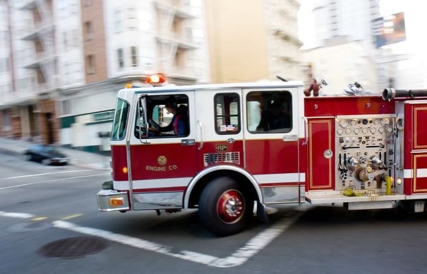 A red and white fire truck is speeding along an urban street, navigating a corner. The background shows blurred buildings, indicating motion. The truck has visible equipment and a lit-up emergency light on top.