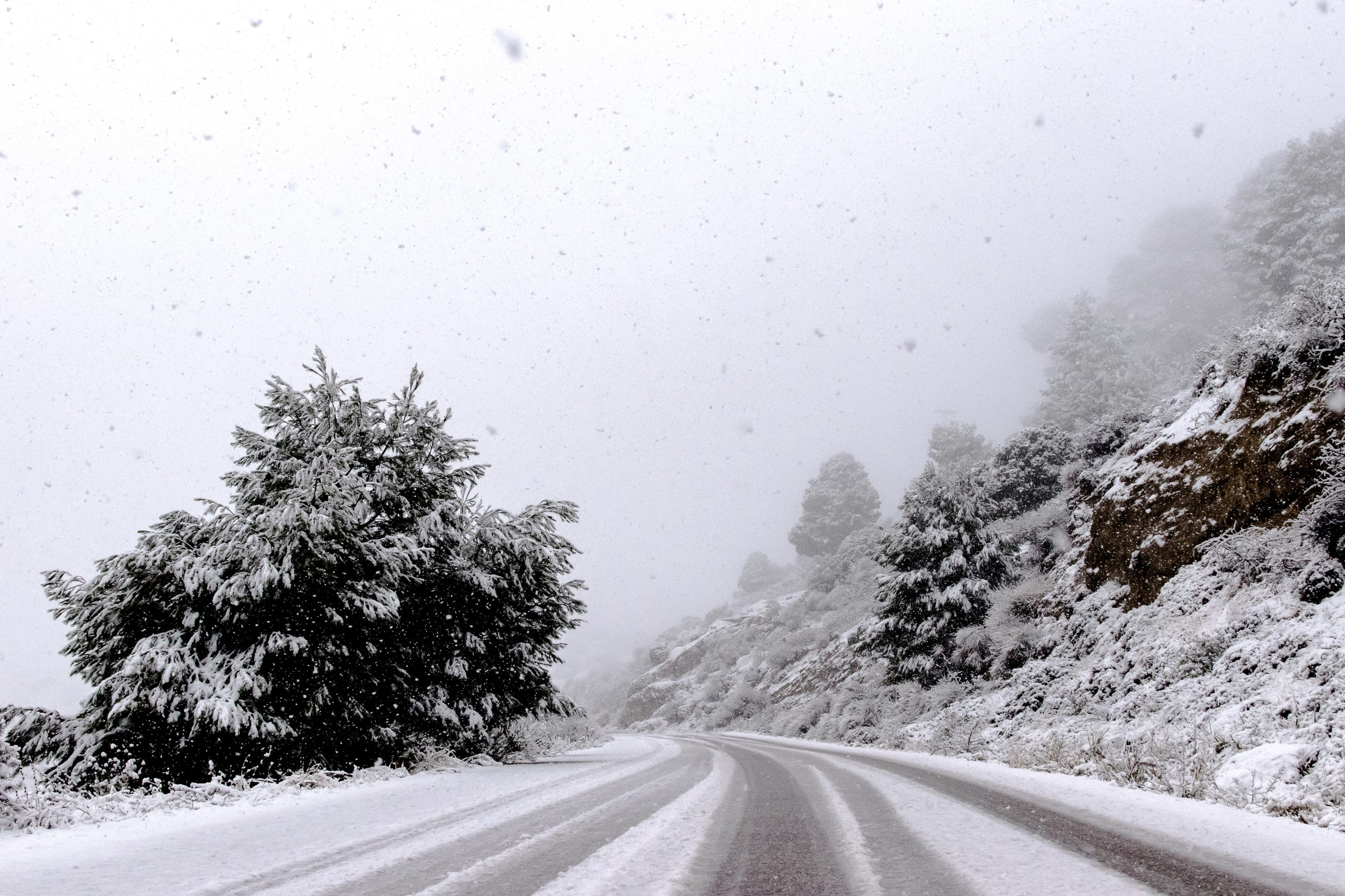 A winding road, blanketed in snow and ice, meanders through the mountainous landscape. Snow-covered trees line both sides of the road, while snow gently falls, creating a misty, wintry scene.