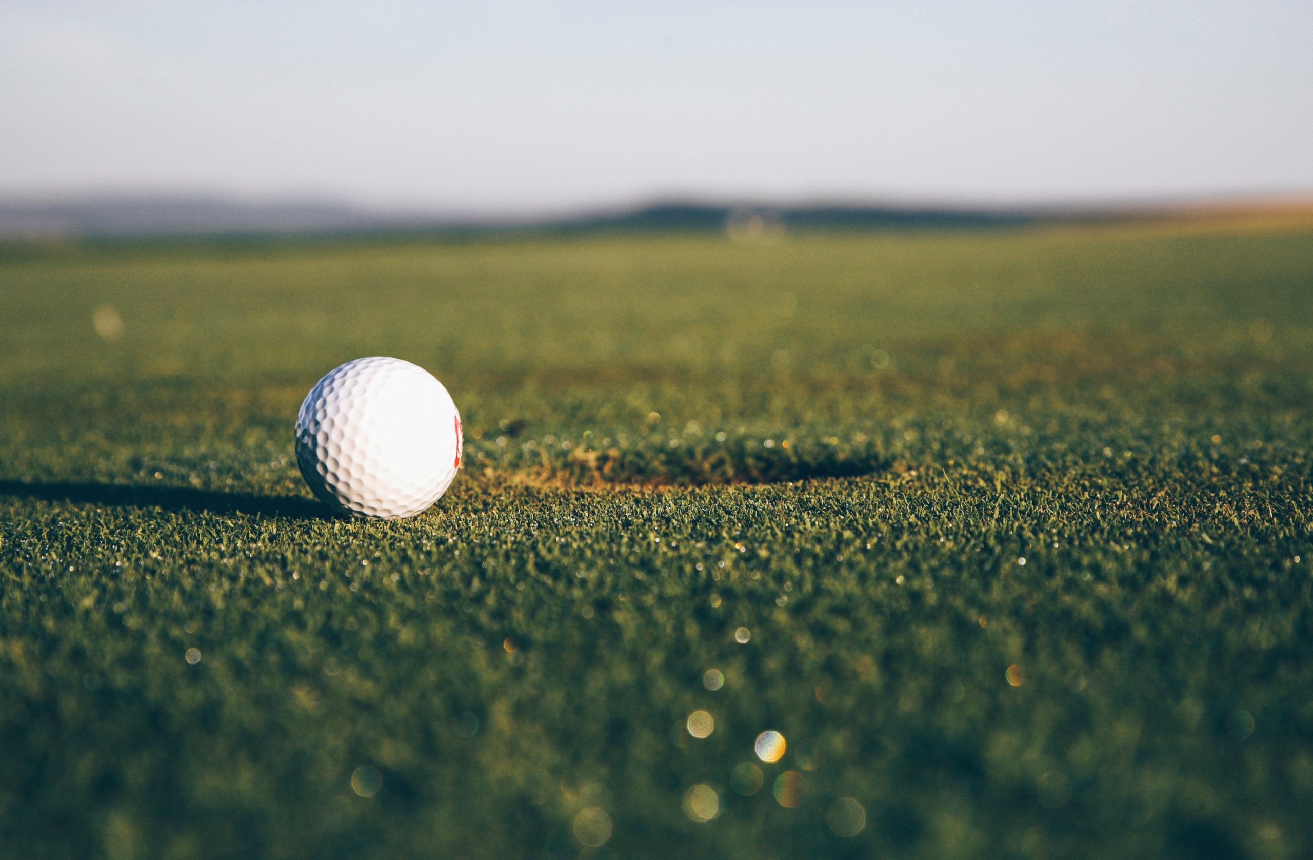 A close-up of a golf ball lies on the green grass near a hole on a golf course, undisturbed by the usual hustle of players or potential golf cart accidents. The background is a clear blue sky and a blurred landscape.