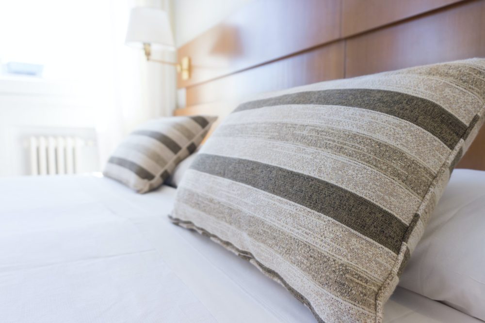 Two striped pillows rest on a neatly made bed with a white sheet. The wooden headboard and wall-mounted lamp evoke the comforting simplicity of a hotel room, without any hint of the usual accidents or mishaps often overheard in bustling hotel corridors.