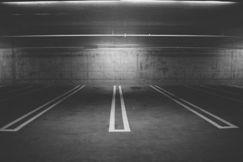 In the dimly lit, black and white image of an empty parking garage, white lines precisely mark the parking spaces against concrete walls and ceiling. This stark setting evokes contemplation: what should you do after a parking lot accident in New Jersey?