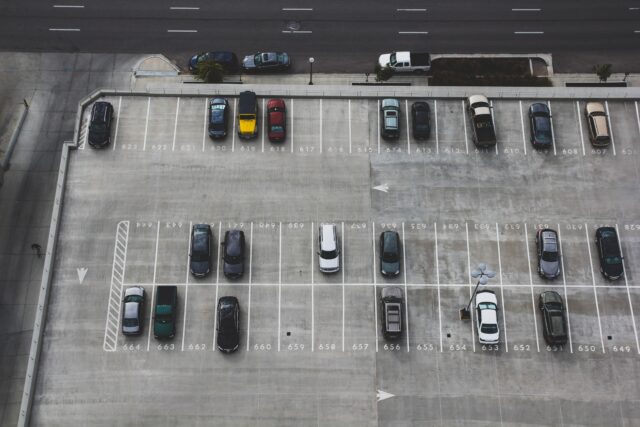 An overhead view shows a parking lot with two rows of parked vehicles on a concrete surface, meticulously marked by white lines and numbers designating each space. A road with several lanes runs parallel to the lot at the top of the image, where, thankfully, no parking lot accident attorneys are needed today.