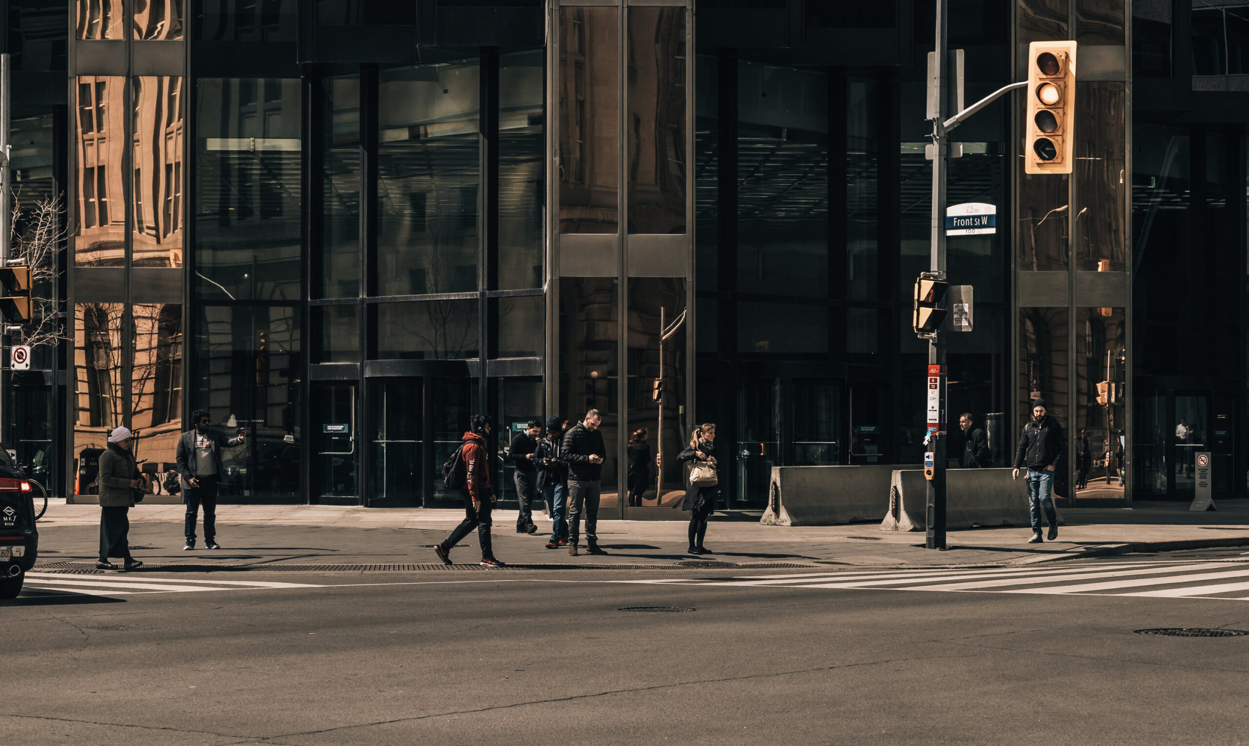 A group of people, possibly unaware of nearby pedestrian accident attorneys, stand at a crosswalk on a city street corner. The building behind them has large glass windows reflecting the opposite structure. Traffic lights and a street sign are visible, and the area appears urban and busy.