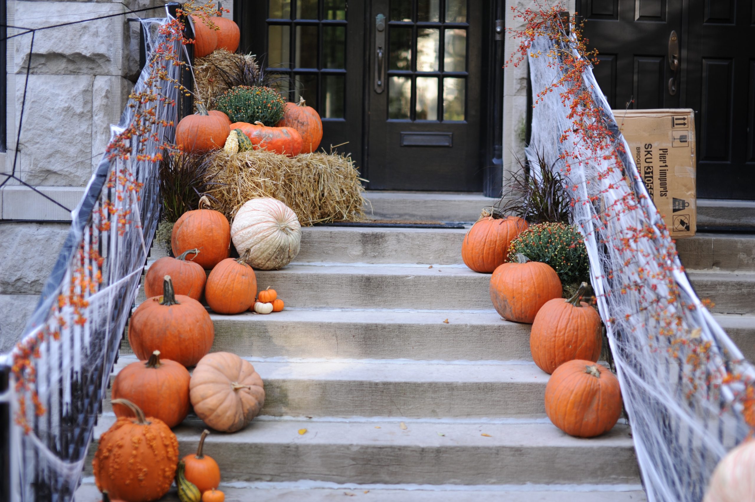 A staircase decorated with pumpkins and hay bales leads to a double-door entrance, perfect for New Jersey trick-or-treating. Both sides of the stairs are lined with orange and white pumpkins, and dried plants. A cardboard box is on the right side near the door.