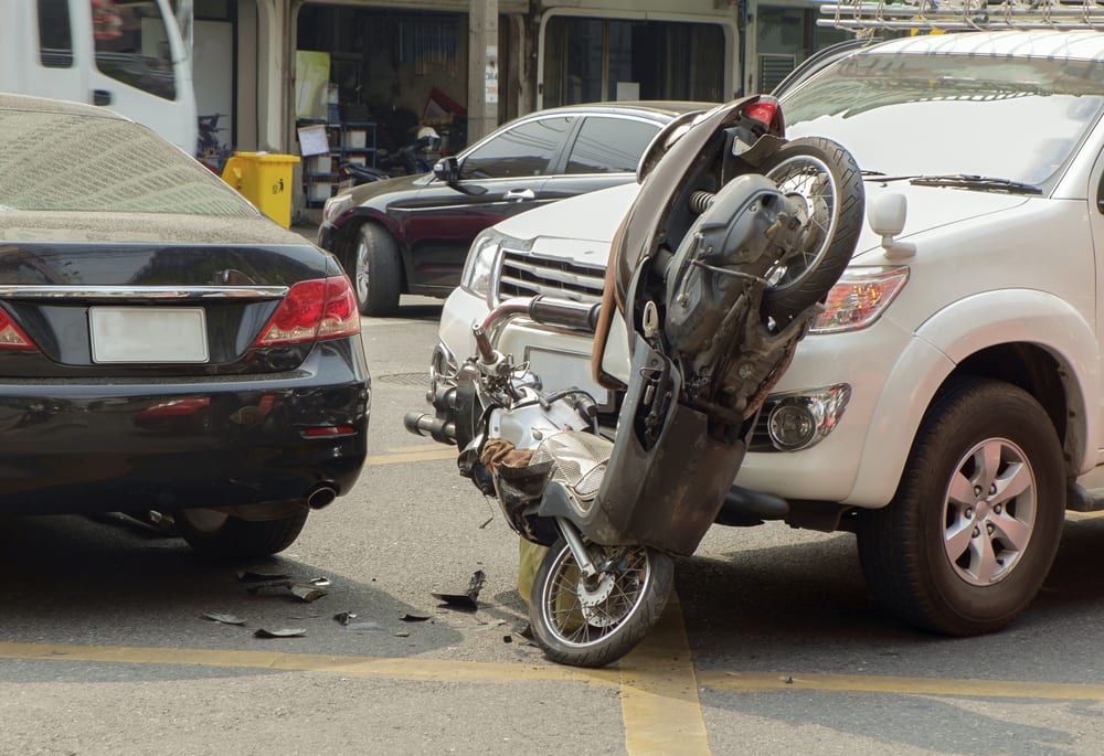 A traffic accident scene showing a motorcycle wedged between two cars, a white SUV and a black sedan. The motorcycle is positioned vertically against the white SUV, with visible damage and debris scattered on the road.