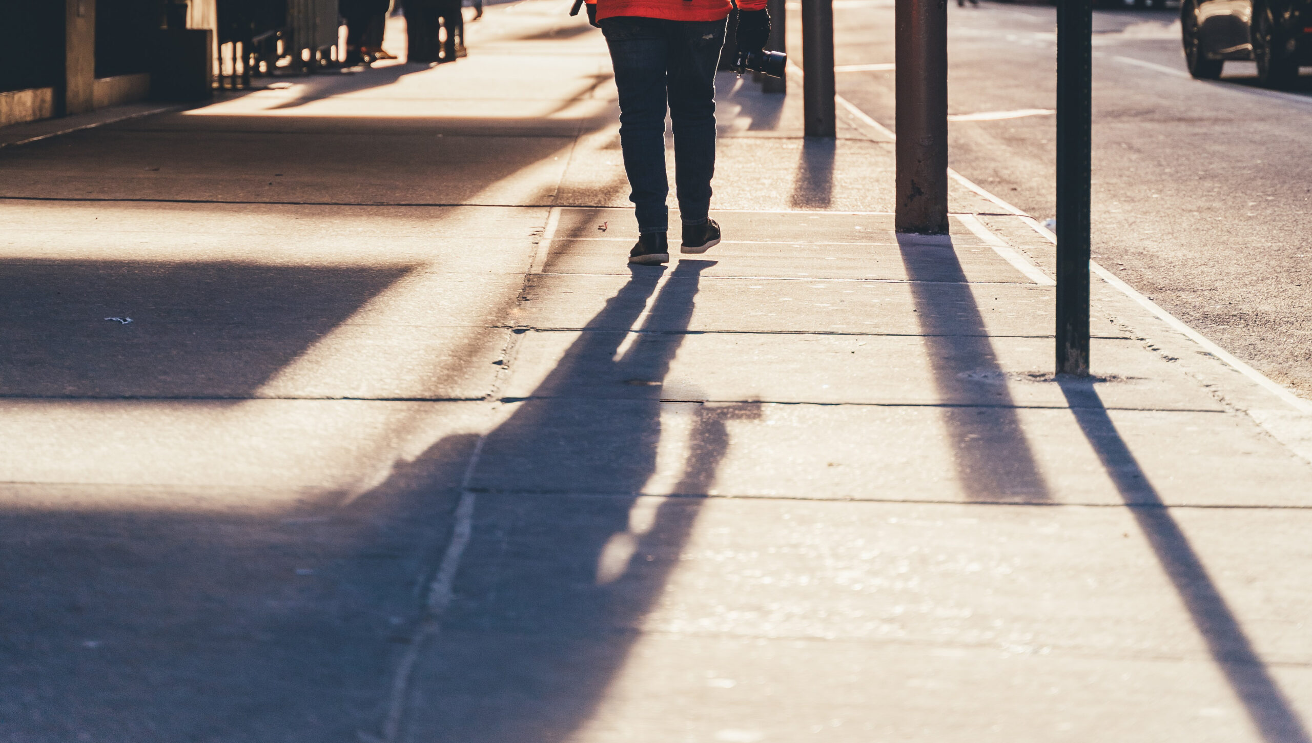 A person walking on a sunlit sidewalk, casting a long shadow. They are wearing dark pants and a red top as distinct lines and textures appear on the pavement. The scene evokes a moment where even Sidewalk Accident Attorneys would pause to admire the warm, elongated reflections as the sun begins to set.