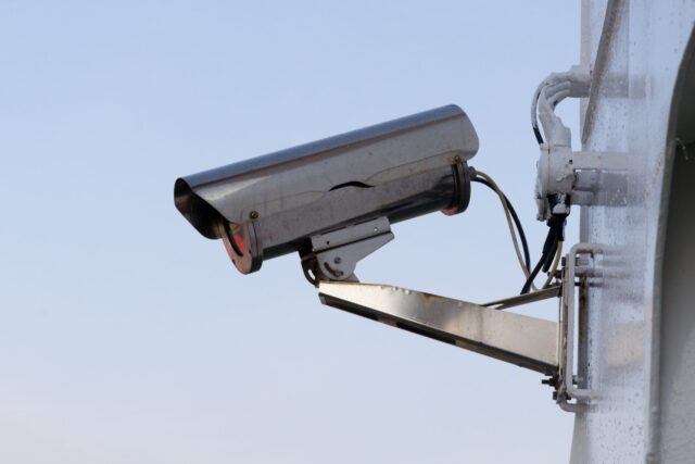 A metal security camera mounted on a white wall at a New Jersey property, captured against a clear blue sky. Despite its presence, the sense of negligence is heightened by the poorly secured, visible cables connecting it to the wall.