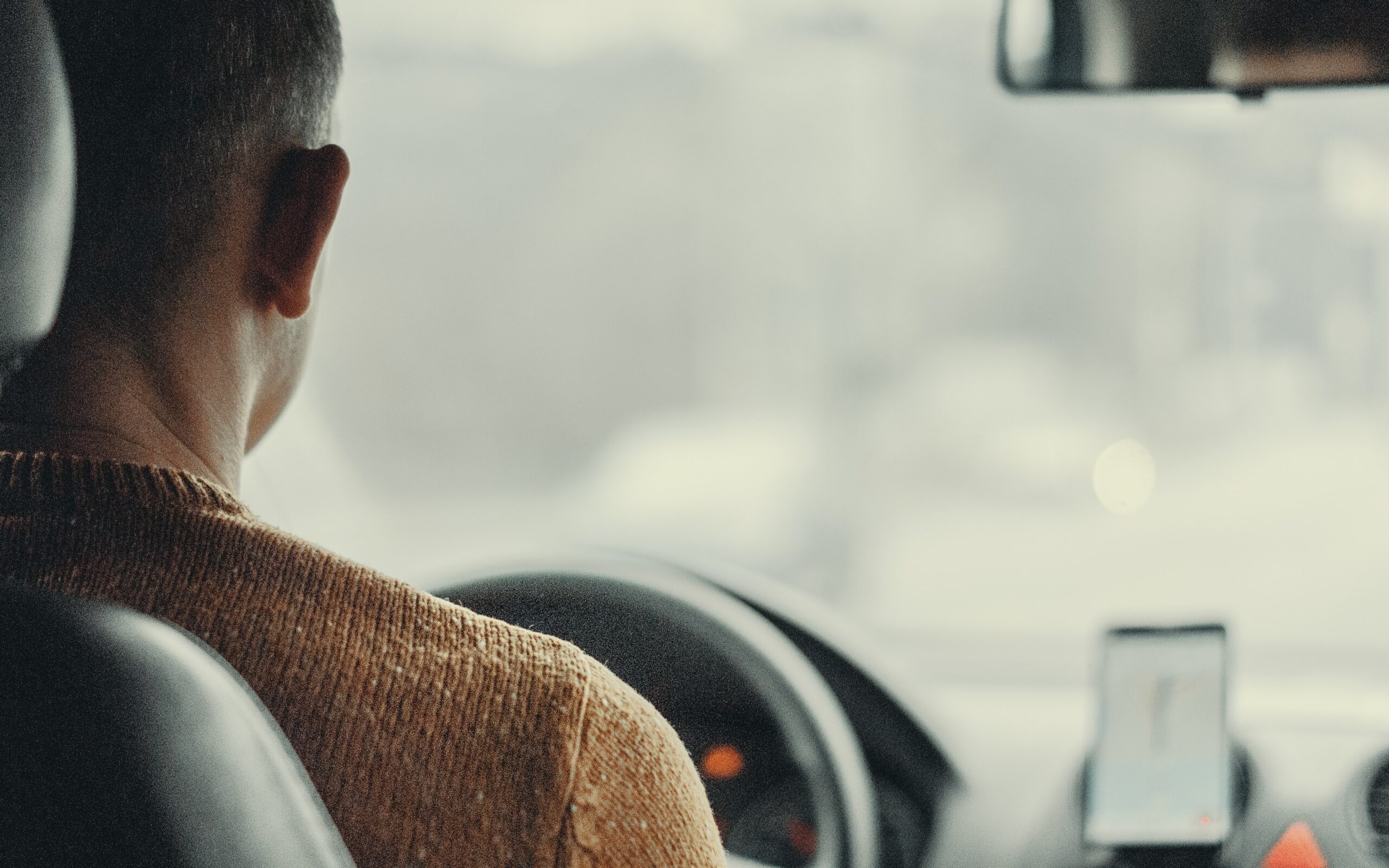 A person driving a car, viewed from the back seat. The driver, perhaps an Uber or Lyft operator, is wearing a brown sweater. A smartphone with a navigation app is mounted on the dashboard. The outside scenery is blurred, suggesting motion and highlighting the importance of safety and accident awareness.