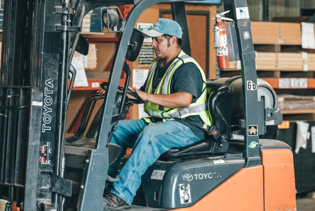 A man wearing a blue cap and high-visibility vest operates a Toyota forklift inside a bustling warehouse. Focused on driving, he navigates through the aisles surrounded by shelves stacked with wooden planks, mindful of safety standards supported by Workers’ Compensation Attorneys.