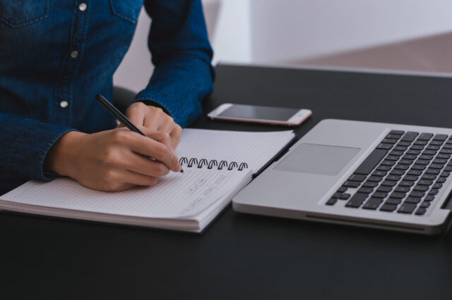 A person in a denim shirt is jotting down notes in a spiral notebook with a pen, while a laptop and smartphone rest on the black tabletop nearby.