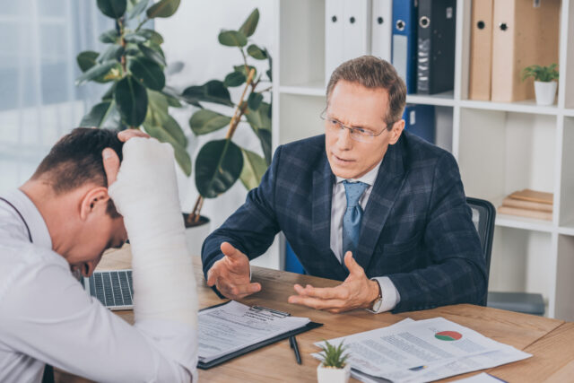 A man with a bandaged arm sits at a desk, looking distressed, discussing his workers' compensation with another man in a suit who gestures as he speaks. Papers and a small plant are on the desk, and shelves with binders are in the background.