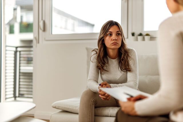 A woman seated on a couch listens attentively to another person whose back is turned to the camera, as if discussing psychological injuries. She is wearing a light-colored sweater and khaki pants in a bright room with large windows and potted plants in the background.